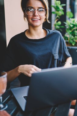 Gelişmiş netbook aracılığıyla yeni ilginç web sitelerini ararken kameraya bakan neşeli kız öğrencinin portresi, görüş düzeltmesi için gözlüklü gülümseyen hippi kız teknolojiyi kullanarak