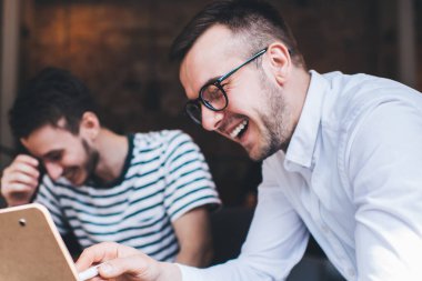 Beyaz gömlekli, gözlüklü, elinde not panosu olan ve kafede iş arkadaşıyla gülen neşeli bir genç.