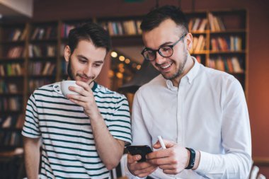 Akıllı telefonları karıştıran ve sıcak içecekler içen neşeli gençler kütüphanedeki serbest meslek sırasında masaya yaslanıp dinleniyorlar.