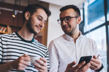 Modern kafede akıllı telefonları karıştırırken ve kahve içerken sıradan giyinmiş, sohbet eden pozitif gülümseyen genç adamlar.
