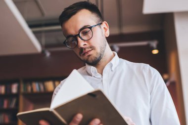 Düşük açılı, gözlüklü, sayfaları çeviren ve halk kütüphanesinin bulanık arka planında durup kitap okuyan erkek.