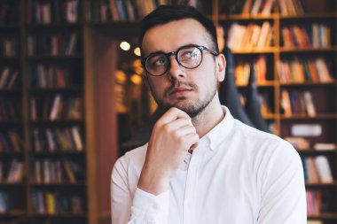 Hipster kahve dükkanında masaya yaslanıp kameraya bakarken çenesine dokunan tişörtlü ve gözlüklü modern bir genç.