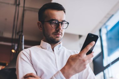 Çalışma sırasında kütüphanenin bulanık arka planında dururken, beyaz gömlekli, kolları katlayan ve akıllı telefonları karıştıran konsantre yöneticiden