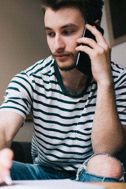 Çizgili tişörtlü ve kot pantolonlu yakışıklı adam evde dizüstü bilgisayarla oturuyor ve akıllı telefondan notlar alırken ortağını dinliyor.