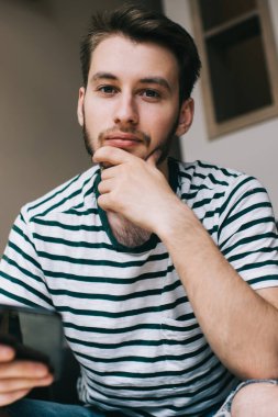 Çizgili tişörtlü, sakallı, akıllı telefon tutan, koltukta oturan ve kameraya bakan düşünceli genç bir adam.