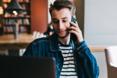 Modern kafede dizüstü bilgisayarda sörf yaparken masada oturup telefonla konuşan serbest yakışıklı, modern bir adam.