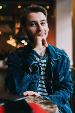 Kot ceketli, kendine güveni tam genç bir adam masaya yaslanmış kafeteryada uzaktan kameraya bakarken dizüstü bilgisayar kullanıyor.