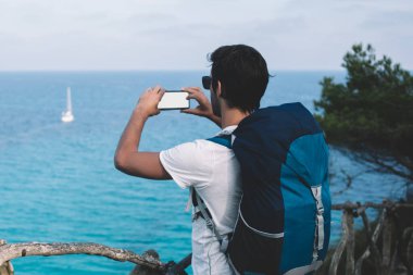 Sırt çantalı meçhul erkek turistin Menorca Adası 'ndaki boş ekran dizüstü bilgisayarında barışçıl mavi okyanusun fotoğrafını çekerken görüntüsü.
