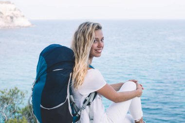 Dertsiz sırt çantalı gezgin, macera yolculuğunda kameraya bakan Menorca 'nın manzara manzarasından keyif alan, turistik sırt çantasıyla gezen neşeli kadın portresi.