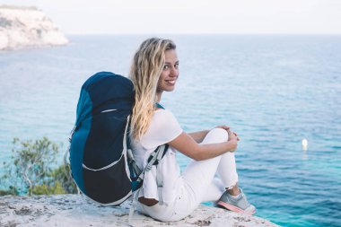 Dertsiz sırt çantalı gezgin, macera yolculuğunda kameraya bakan Menorca 'nın manzara manzarasından keyif alan, turistik sırt çantasıyla gezen neşeli kadın portresi.