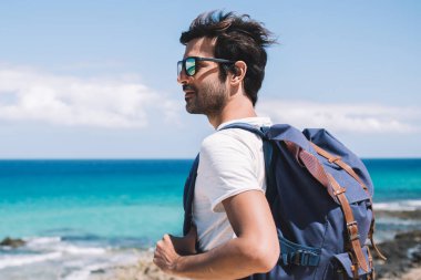 Rüzgârlı, saçı rüzgarlı neşeli gezgin adam deniz kenarında durup bakarken gülümseyen ve hayranlık duyan cennet gibi deniz manzarası