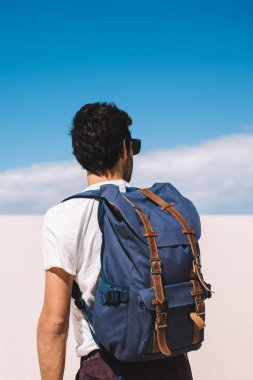 Güneş gözlüklü, sırt çantalı, kum tepeleri boyunca güneşin altında gezinen ve çöl manzarasına bakan erkek turistin arkası.