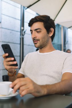 Düşük açılı, rahat, serbest giyinen, perdeye odaklanan ve akıllı telefonu faiziyle kullanan, modern kahve dükkanının terasında bir fincan lezzetli sıcak içecekle tek başına oturan bir erkek.