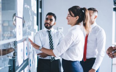 Konferans odasındaki modern cam panoya yapıştırılmış bilgi grafikleri üzerinde çalışan çeşitli ofis çalışanları, mutlu ortaklar brifingi ve iletişim...