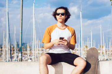 Genç, kıvırcık saçlı, güneş gözlüklü ve sıradan kıyafetli bir adam. Barcelona 'daki güzel rıhtımda kayalık çitlerde otururken cep telefonuyla sohbet ediyor.