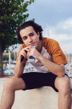 Tatlı kıvırcık saçlı adam kayalık çitlerde dinleniyor gülümsüyor ve kameraya bakıyor Barcelona 'da güneşli bir günde dirseğini dizlerine dayayıp kameraya bakıyor