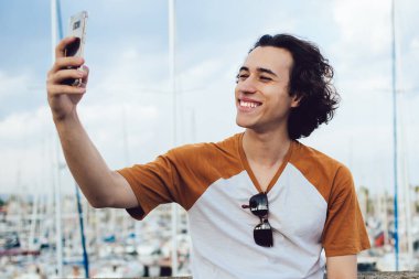 Neşeli kıvırcık saçlı erkek gezgin gülümseyerek gülümserken cep telefonuyla barselona arka planında renkli iskele olan çitlere yaslanarak selfie çekiyor.