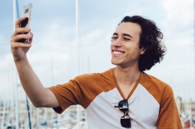 Neşeli kıvırcık saçlı erkek gezgin gülümseyerek gülümserken cep telefonuyla barselona arka planında renkli iskele olan çitlere yaslanarak selfie çekiyor.