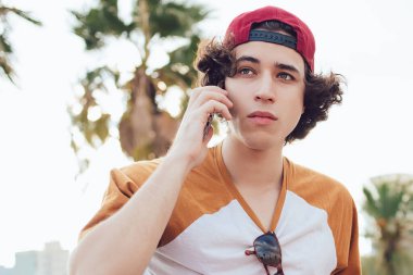 Siyah saçlı, günlük kıyafetleri ve şapkası olan, cep telefonuyla konuşan ve Barselona 'da bulanık arka planda tropik avuçlarla bakan adamın yanından.