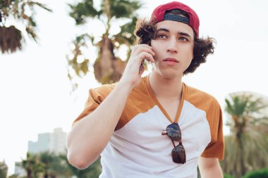 Siyah saçlı, günlük kıyafetleri ve şapkası olan, cep telefonuyla konuşan ve Barselona 'da bulanık arka planda tropik avuçlarla bakan adamın yanından.