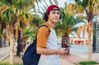 Sıradan giyinen, sırt çantalı, şapkalı, gülümseyen ve elinde cep telefonuyla Barcelona 'da egzotik avuçlarıyla kameraya bakan içerikli erkek hipsterin yan görüntüsü.