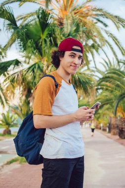 Sırt çantalı kaygısız turist, güneşli bir sokakta palmiye ağaçlarıyla dikiliyor ve yaz tatillerinde sıcak bir ülkeyi geziyor. Dijital akıllı telefonuyla gezen genç gezgin.