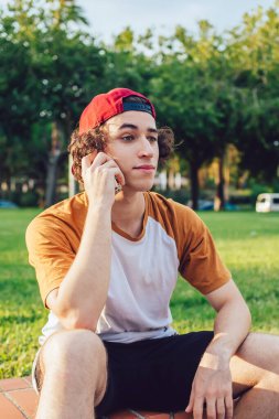 Kıvırcık saçlı, günlük kıyafetleri ve kırmızı şapkası olan genç bir adam. Tuğla çitin üzerinde oturuyor ve Barcelona 'da güneşli bir günde cep telefonuyla sohbet ediyor.