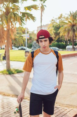 Spor kıyafetli bir erkek, güneş ışığında palmiye ağaçlarıyla boş bir yolda kaykay yaparken kameraya bakıyor ve bir elini cebine sokuyor.