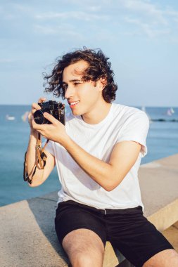 Kıvırcık saçlı, gülümseyen adam deniz kıyısında taş çitlerin üzerinde oturuyor ve kamerayla gökyüzüne bakarken yüzünü çeviriyor.