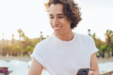 Beyaz gömlekli, siyah pantolonlu, çitlere yaslanmış gülümseyen genç bir adam. Güneşli Barcelona 'da telefonu elinde tutan dişlek bir gülümsemeyle başka tarafa bakıyor.