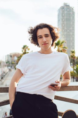 Barcelona 'da cep telefonuyla ahşap çitlere yaslanırken kameraya bakan, modaya uygun giyinen iri yapılı genç adam.