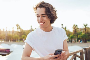 Beyaz gömlekli, siyah pantolonlu, çitlere yaslanmış gülümseyen genç bir adam. Güneşli Barcelona 'da telefonu elinde tutan dişlek bir gülümsemeyle başka tarafa bakıyor.