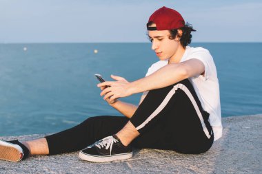 Kırmızı şapkalı düşünceli genç adam gün batımında temiz suya ve bulutlu gökyüzüne karşı taş çitlerde serinlerken akıllı telefonuyla mesaj atıyor.