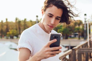 Yakışıklı, kıvırcık saçlı, gündelik giyinmiş, Barcelona 'da ahşap çitlere yaslanırken cep telefonu izleyen bir adam.