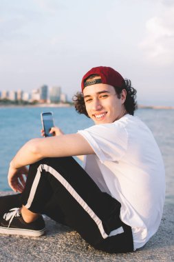 Beyaz tişörtlü hippi kıyı şeridinin çitlerinde cep telefonu kullanarak kameraya gülümseyen ve fotoğraf çeken adamın yan görüntüsü.