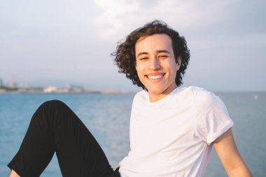 Beyaz tişörtlü genç adam taştan çitlerin üzerinde oturuyor şehir ve deniz manzarasına karşı kameraya gülümserken güneşleniyor.