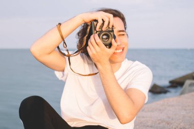 Deniz kenarındaki arka planda oturan yeniyetme erkek, taş çitlerde oturuyor ve günbatımının fotoğraflarını çekiyor.