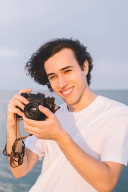 Deniz ve bulutlu gökyüzünün arka planında duran mutlu genç adam gün batımının fotoğrafını çekiyor ve yaz tatilinin tadını çıkarırken gülümsüyor.