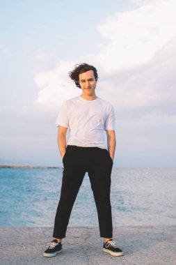 Neşeli hippi adam, gündelik giysiler içinde taş çitin üzerinde el ele tutuşmuş ve denizde vakit geçirirken kameraya bakıyor.