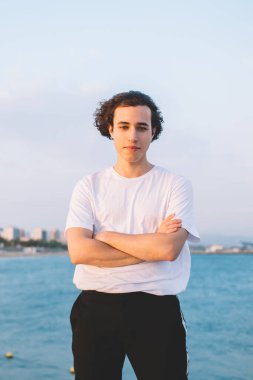 Beyaz tişörtlü düşünceli bir adam taş çitin üzerinde el ele tutuşup kameraya bakarken hayal kuruyor ve gezinin tadını çıkarıyor.