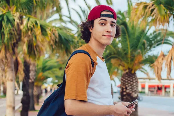 Sıradan giyinen, sırt çantalı, şapkalı, gülümseyen ve elinde cep telefonuyla Barcelona 'da egzotik avuçlarıyla kameraya bakan içerikli erkek hipsterin yan görüntüsü.