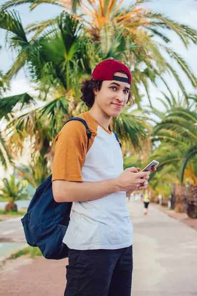 Sırt çantalı kaygısız turist, güneşli bir sokakta palmiye ağaçlarıyla dikiliyor ve yaz tatillerinde sıcak bir ülkeyi geziyor. Dijital akıllı telefonuyla gezen genç gezgin.
