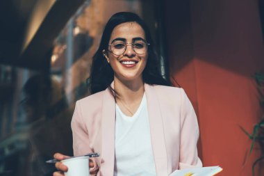 Eğitim kitabı ve kahvesi olan zeki bir bayan öğrenci kameraya gülümseyecek. Optik gözlüklü neşeli İspanyol kadının yarım boy portresi kaldırımda bilgi öğreniyor.