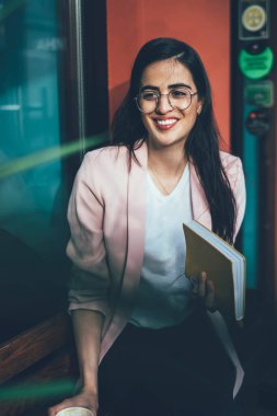 Görme koruması için gözlüklü mutlu İspanyol bayan öğrenci edebiyat hobisi için zaman harcıyor, şehir ortamında çok satan kitabı olan neşeli Gürcü kadın gülümsüyor.