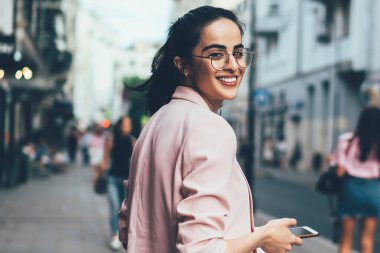 Tek başına seyahat ederken kameraya gülümseyen cep telefonu cihazlı neşeli kadın turistin yarım boy portresi optik gözlüklü hippi kız elinde akıllı telefonuyla kentselliğe poz veriyor.