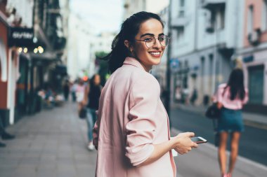 Tek başına seyahat ederken kameraya gülümseyen cep telefonu cihazlı neşeli kadın turistin yarım boy portresi optik gözlüklü hippi kız elinde akıllı telefonuyla kentselliğe poz veriyor.
