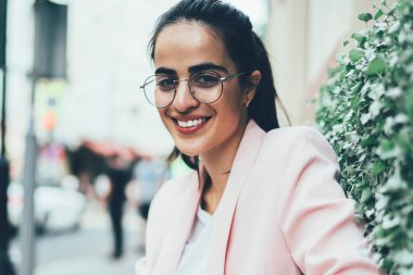 Şehir bankında dinlenirken kameraya gülümseyen moda gözlüklü komik kadın blogcu portresi, akıllı telefon teknolojisiyle şehirde keyif çatan mutlu İspanyol kadın.