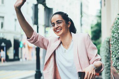 Görme düzeltmesi için klasik gözlüklü neşeli İspanyol kadın şehirde beklenmedik bir canlı buluşma mutlu 20 'li yaşlarda elinde cep telefonu zımbırtıları ve tezgahta arkadaşını beklerken el sallıyor.