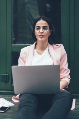 Laptop bilgisayarlı düşünceli bayan öğrenci araştırma sırasında grafik grafikleri hakkında düşünüyor ve şehir ortamında öğreniyor, gözlüklü İspanyol kadın bilgi grafikleri üzerinde uzaktan çalışıyor.