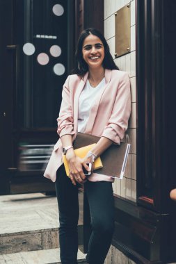 Moda kıyafetler giymiş, elinde eğitim ekipmanları olan neşeli hippi kız şehir binasının yanında duruyor ve gülümsüyor, eskiz defteri ve not defteriyle şehir sokaklarına gülüyor.